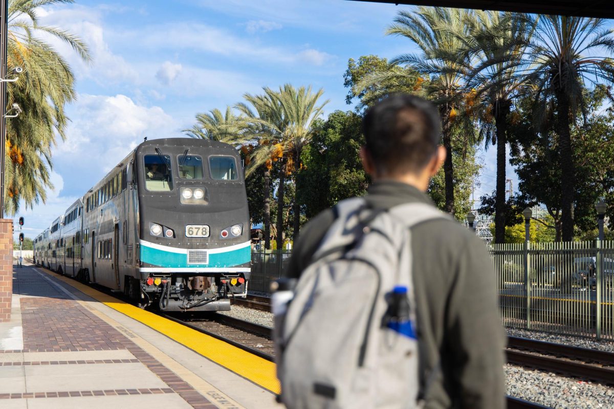 A train approaching a station in a beach area. A male student with a grey backpack is watching its approach.